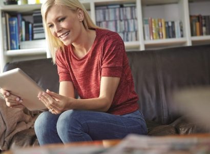 Vrouw zit op bank en kijkt lachend naar de tablet die zij in haar handen houdt.